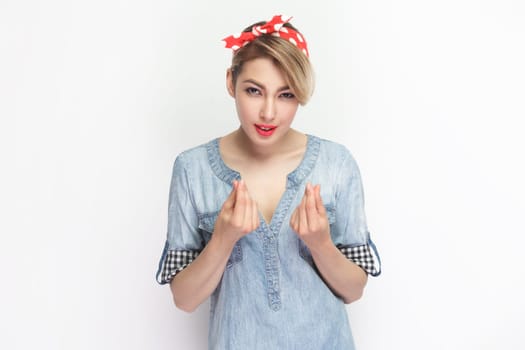 Portrait of attractive beautiful blonde woman wearing blue denim shirt and red headband standing showing money gesture with fingers. Indoor studio shot isolated on gray background.