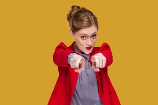 Portrait of amazed positive optimistic woman with red lips standing pointing finger at camera, selecting you, wearing red jacket. Indoor studio shot isolated on yellow background.