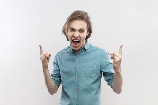 Portrait of crazy excited young man having fun at rock festival, showing rock and roll gesture, screaming, yelling, wearing blue shirt. Indoor studio shot isolated on gray background.