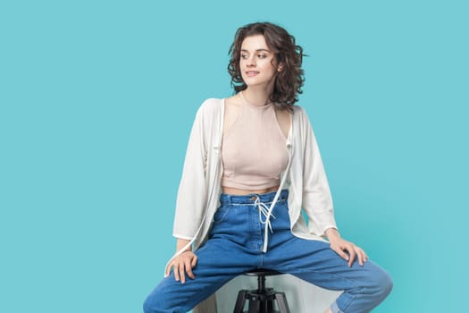 Portrait of positive cheerful beautiful young adult woman sitting on chair and posing like fashion model and looking away with happy smile. Indoor studio shot isolated on blue background.