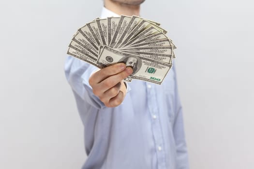 Portrait of unknown anonymous man standing holding out big fan of dollar banknotes, showing his salary, wearing light blue shirt. Indoor studio shot isolated on gray background.