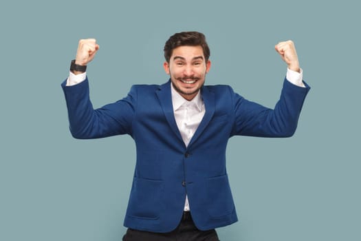 Portrait of self-confident happy satisfied man with mustache standing with raised arms, showing his power, wearing official style suit. Indoor studio shot isolated on light blue background.