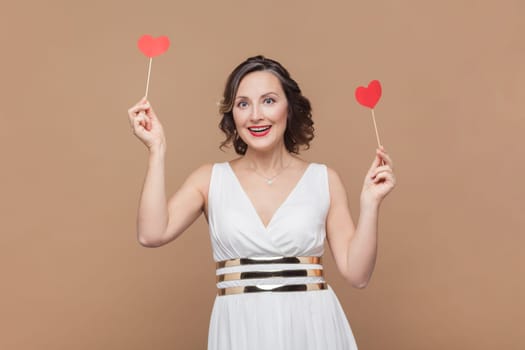 Portrait of excited positive middle aged woman with wavy hair holding little red hearts, smiling happily, celebrating, wearing white dress. Indoor studio shot isolated on light brown background.
