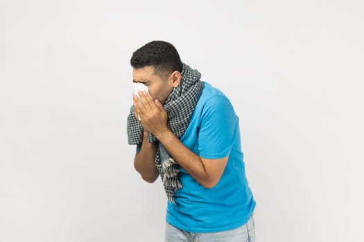 Portrait of ill sick unshaven man wearing blue T- shirt and wrapped in warm scarf standing sneezing, having runny nose, has grippe. Indoor studio shot isolated on gray background.