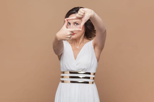 Portrait of middle aged woman with wavy hair makes frame gesture over head, searches perfect angle, makes picture, wearing white dress. Indoor studio shot isolated on light brown background.