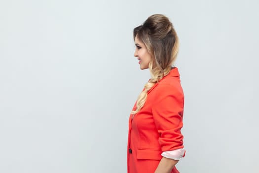 Side view portrait of smiling attractive positive optimistic blonde woman standing looking ahead, expressing positiveness, wearing red jacket. Indoor studio shot isolated on gray background.