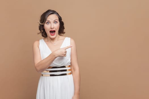 Portrait of happy joyful middle aged woman with wavy hair pointing away at copy space for advertisement, wearing white dress. Indoor studio shot isolated on light brown background.