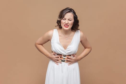 Portrait of unhealthy sick middle aged woman with wavy hair standing, holding her belly with hands, stomach cramps, wearing white dress. Indoor studio shot isolated on light brown background.