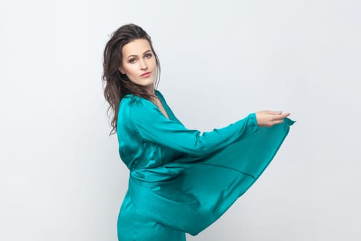 Side view portrait of elegant attractive beautiful young adult brunette woman with wavy hair, looking at camera, wearing green dress. Indoor studio shot isolated on gray background.