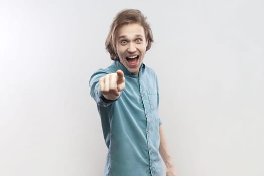 Portrait of surprised astonished shocked young man standing pointing at camera, selecting you, expressing positive emotions, wearing blue shirt. Indoor studio shot isolated on gray background.