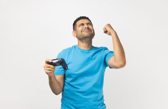 Cheerful unshaven man gamer wearing blue T- shirt standing playing video games, holding joystick and clenched fist, celebrating his winning. Indoor studio shot isolated on gray background.