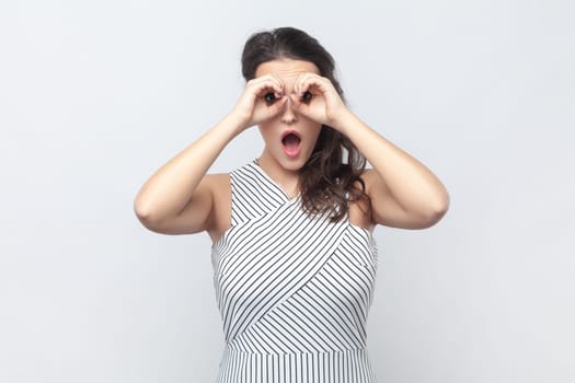Portrait of surprised interested brunette woman making binoculars gesture with hands and looking amazed at camera, wearing striped dress. Indoor studio shot isolated on gray background.