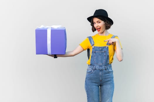 Portrait of attractive smiling hipster woman in blue denim overalls, yellow T-shirt and black hat, holding and pointing at present box. Indoor studio shot isolated on gray background.