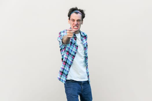 Furious angry man looses temper, screams and points at you, blames someone and expresses negative emotions, wearing checkered shirt and headband. Indoor studio shot isolated on gray background.