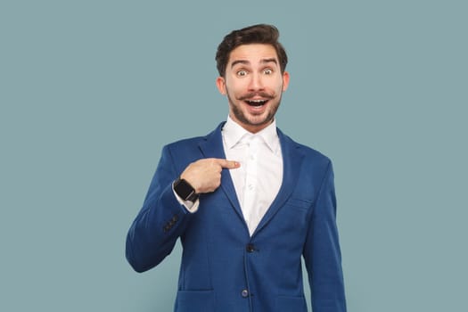 Portrait of excited amazed handsome man with mustache standing pointing at himself with surprised face, wearing official style suit. Indoor studio shot isolated on light blue background.