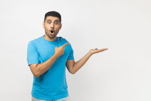 Portrait of amazed excited surprised astonished unshaven man wearing blue T- shirt standing presenting advertisement area on his palm, mockup. Indoor studio shot isolated on gray background.