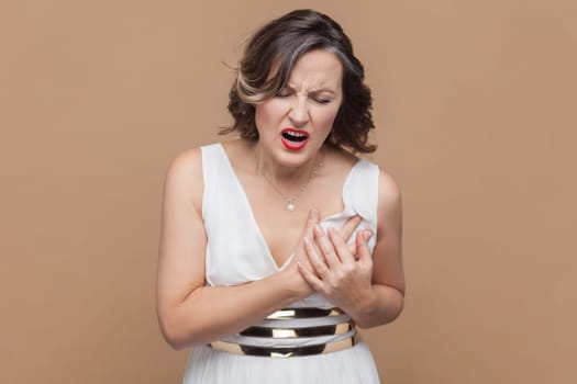 Middle aged woman with wavy hair holding hands on chest feeling acute pain in chest, risk of stroke, heart attack, wearing white dress. Indoor studio shot isolated on light brown background.