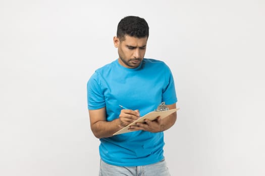 Portrait of serious concentrated unshaven man wearing blue T- shirt standing with clipboard, writing something on paper, making to do list. Indoor studio shot isolated on gray background.