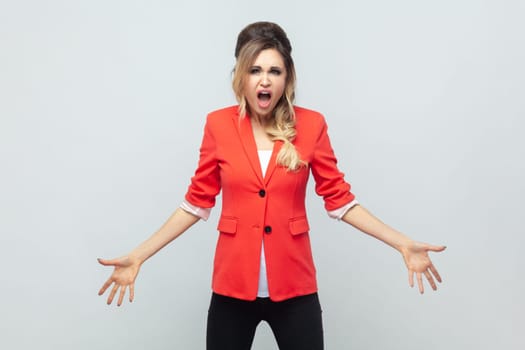 Portrait of frustrated irritated angry blonde woman standing with spread hands, screaming with hate and anger, negative emotions, wearing red jacket. Indoor studio shot isolated on gray background.