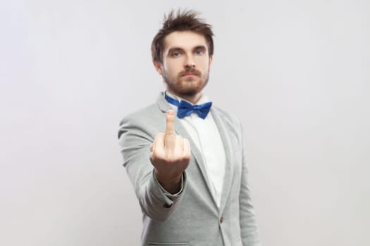 Portrait of rude impolite handsome bearded man standing showing middle finger, arguing with somebody, wearing grey suit and blue bow tie. Indoor studio shot isolated on gray background.