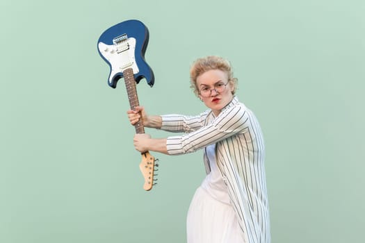 Portrait of angry young adult blonde woman in white striped blouse with eyeglasses standing, holding electric guitar and attacking. Indoor studio shot isolated on light green background.