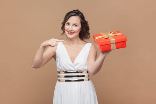 Joyful cheerful middle aged woman with wavy hair pointing at red wrapped present box, congratulating with birthday, wearing white dress. Indoor studio shot isolated on light brown background
