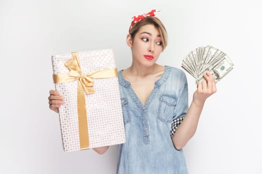 Portrait of confused puzzled uncertain blonde woman wearing blue denim shirt and red headband standing holding present box and dollar banknotes. Indoor studio shot isolated on gray background.