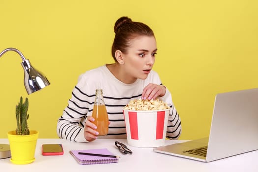 Concentrated woman sitting on workplace and watching interesting movie during break, eating popcorn and drinking beverage. Indoor studio studio shot isolated on yellow background.
