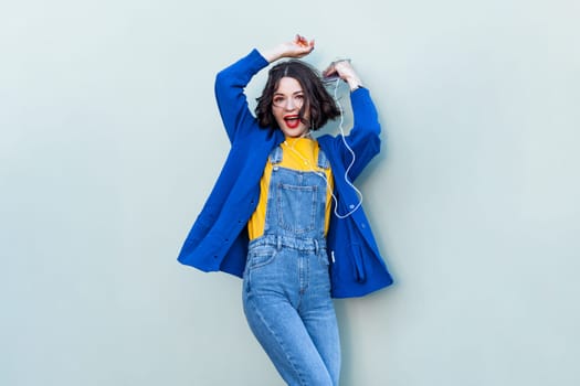 Portrait of happy cheerful hipster woman in denim overalls, yellow T-shirt and blue jacket, listening to music, dancing with raised hands. Indoor studio shot isolated on light green background.