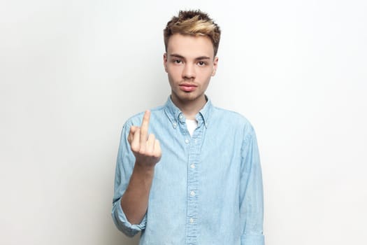 Portrait of rude serious impolite man wearing denim shirt showing middle finger, arguing with somebody, looking at camera with bossy expression. Indoor studio shot isolated on gray background.