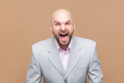 Portrait of angry or shocked bald bearded man standing, looking at camera and screaming, arguing with friend, wearing gray jacket. Indoor studio shot isolated on brown background.
