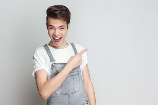 Portrait of young brunette man standing pointing away on copy space, showing amazing promo, gives way or direction, wearing denim overalls. Indoor studio shot isolated on gray background.