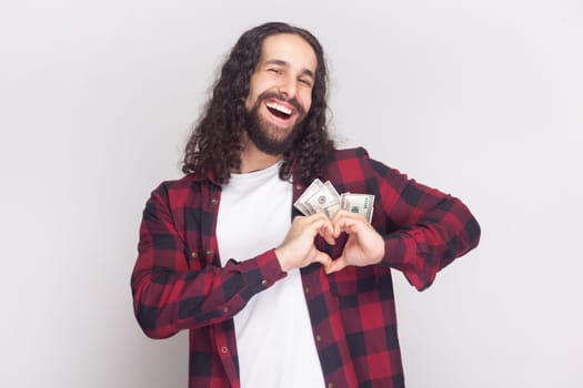 Portrait of smiling joyful delighted man with long curly hair in checkered red shirt holding dollars in pocket, and showing heart shape with hands. Indoor studio shot isolated on gray background.