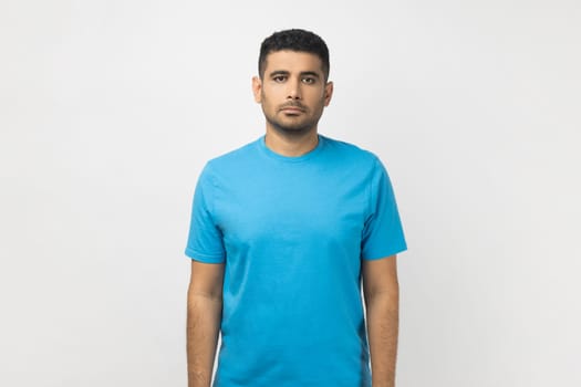 Portrait of serious strict handsome unshaven man wearing blue T- shirt standing looking at camera, having concentrated expression. Indoor studio shot isolated on gray background.