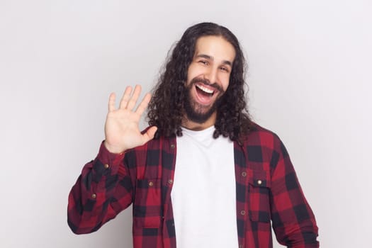 Portrait of friendly-looking bearded man with long curly hair in checkered red shirt saying hi, waving his hand, looking at camera with happy expression. Indoor studio shot isolated on gray background