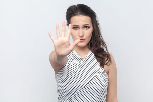 Portrait of serious brunette woman showing stop sign with palm, demonstrates prohibition and restriction, refuses something, wearing striped dress. Indoor studio shot isolated on gray background.