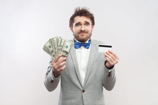 Portrait of sad upset disappointed bearded man showing credit card and big fan of dollar banknotes, wearing grey suit and blue bow tie. Indoor studio shot isolated on gray background.