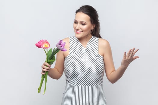 Portrait of amazed happy smiling brunette woman gets bouquet of spring flowers, holding flowers, being glad, pleasant surprise, wearing striped dress. Indoor studio shot isolated on gray background.