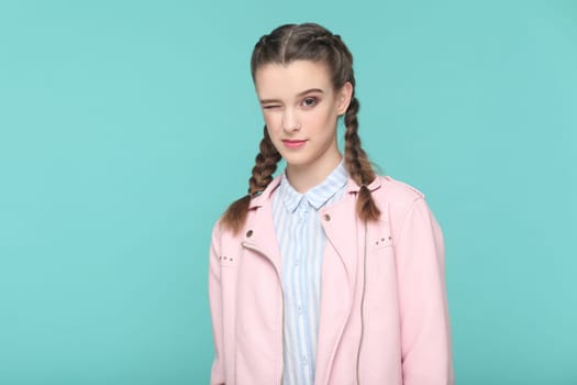 Portrait of playful cheerful teenager girl with braids wearing pink jacket standing looking at camera, winking, flirting with boyfriend. Indoor studio shot isolated on green background.