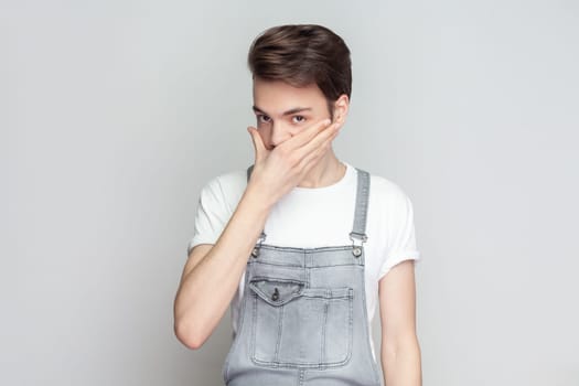 Man standing covering mouth with both hands keeping a secret, doesn't want to spread rumors or some confidential information, wearing denim overalls. Indoor studio shot isolated on gray background.