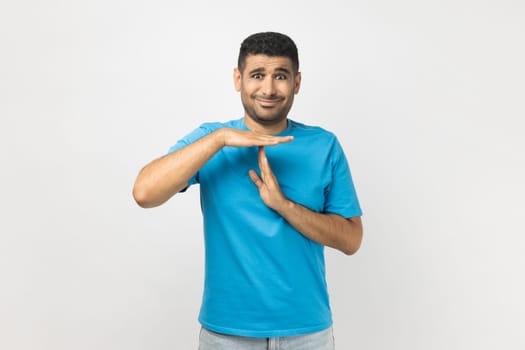 Portrait of attractive young adult unshaven man wearing blue T- shirt standing showing time out gesture with hands, looking at camera. Indoor studio shot isolated on gray background.