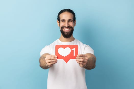 Portrait of man with beard wearing white T-shirt holding out heart like icon of social media and looking at camera with toothy smile. Indoor studio shot isolated on blue background.