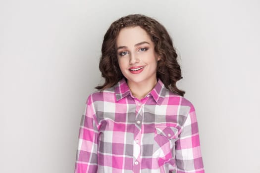 Cheerful woman with curly hair enjoys good day, looks unbothered and happy, gives sincere friendly smile to you, wearing pink checkered shirt. Indoor studio shot isolated on gray background.