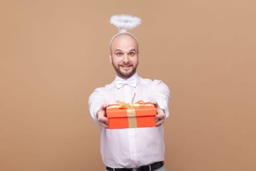 Portrait of friendly bald bearded man with nimb over head, congratulating with Valentines Day, giving present, wearing light pink shirt and bow tie. Indoor studio shot isolated on brown background.