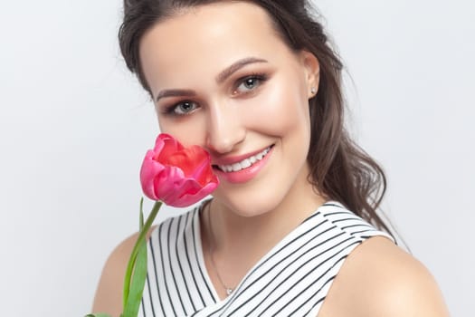 Closeup portrait of romantic smiling joyful brunette woman standing looking at camera, holding pink tulip, enjoying flower, wearing striped dress. Indoor studio shot isolated on gray background.