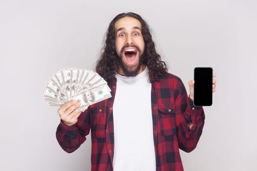 Portrait of amazed excited bearded man with long curly hair in checkered red shirt holding dollar banknotes and cell phone with mockup blank display. Indoor studio shot isolated on gray background.