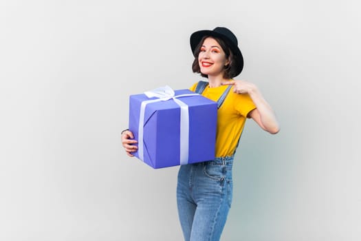 Portrait of smiling pretty hipster woman in blue denim overalls, yellow T-shirt and black hat holding pointing at big present box with gift for birthday. Indoor studio shot isolated on gray background