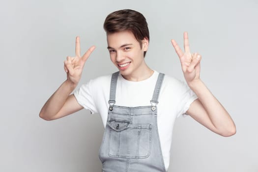 Portrait of young brunette man standing makes peace gesture, shows v sign, looking at camera with toothy smile, wearing denim overalls. Indoor studio shot isolated on gray background.