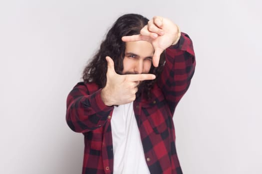 Bearded man with long curly hair in checkered red shirt got inspiration imagines hot to capture interesting shot makes frame gesture smiles gladfully. Indoor studio shot isolated on gray background.
