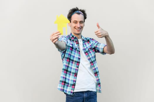 Attractive man holding yellow paper small house and propose you to planning new home to buy, thumb up, wearing blue checkered shirt and headband. Indoor studio shot isolated on gray background.
